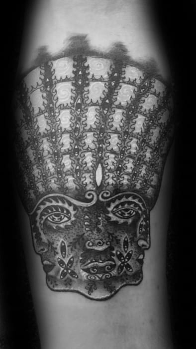 Black and grey Tool-inspired tattoo featuring a symmetrical face with ornate patterns and vertical rows of eye-like shapes extending upward.