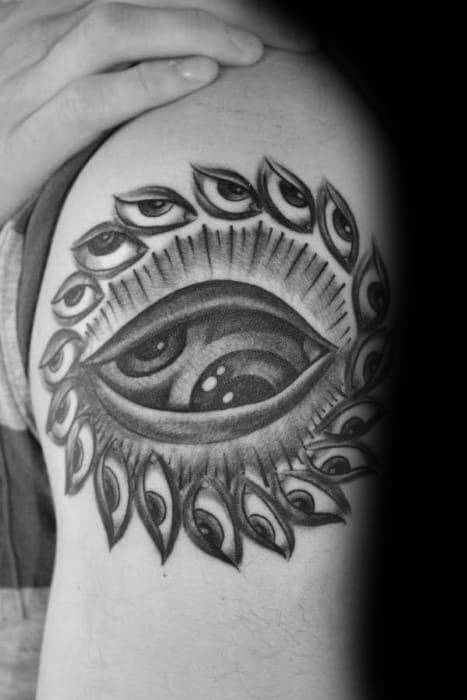 Black and grey Tool-inspired upper arm tattoo of a central eye surrounded by smaller eyes in a circular formation, with bold shading and surreal detail.