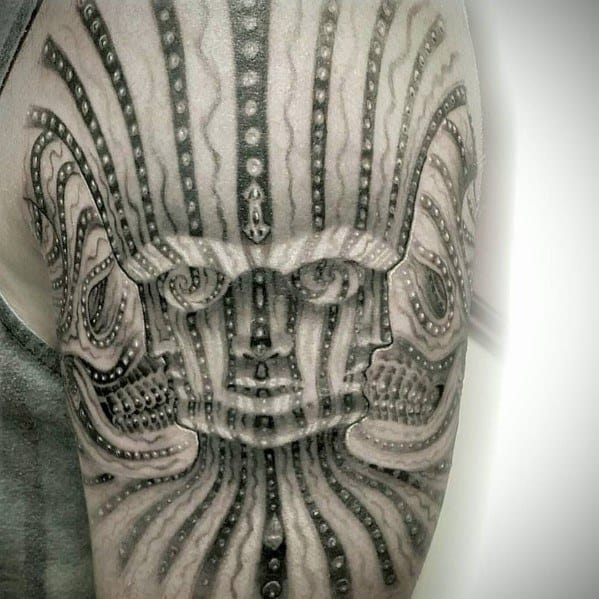 Black and grey Tool-inspired upper arm tattoo featuring a symmetrical, abstract face with dotwork detailing and flowing vertical patterns.