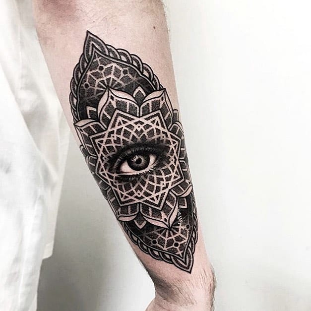 Tattoo of a realistic eye within an intricate mandala pattern, inked in black and grey shading on the forearm.