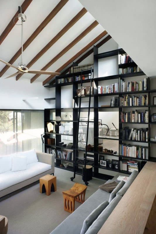Modern living room with high ceiling, exposed beams, large bookcase with ladder, and cozy seating area