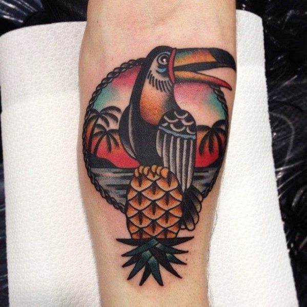 Traditional-style toucan tattoo with a pineapple, tropical sunset, and palm trees, framed in bold lines and vibrant colors on the forearm.
