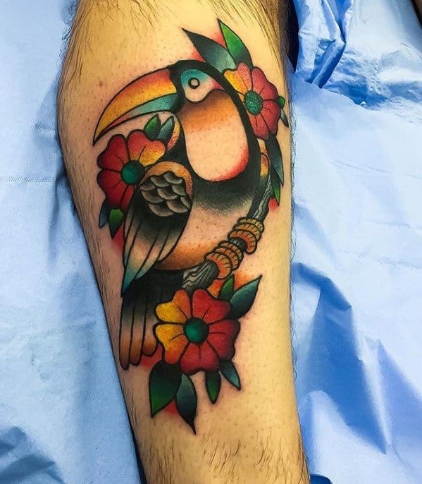 Traditional-style toucan tattoo on the leg with bold black lines, colorful flowers, and vibrant green, red, and yellow shading.