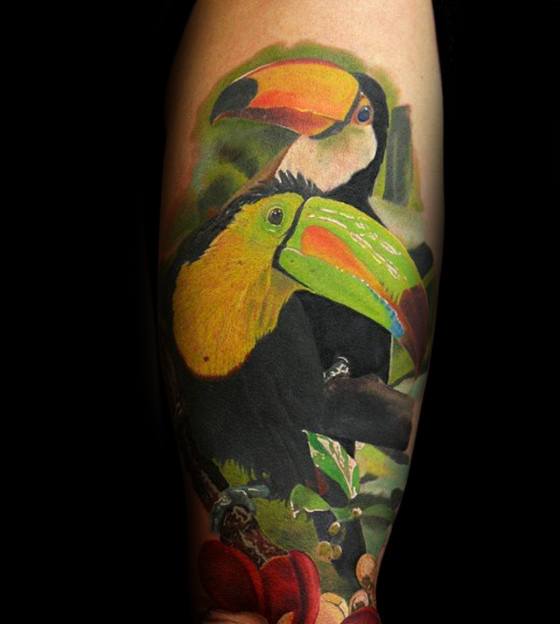 Hyper-realistic tattoo of two toucans with vivid yellow and green beaks, surrounded by detailed jungle foliage on the forearm.