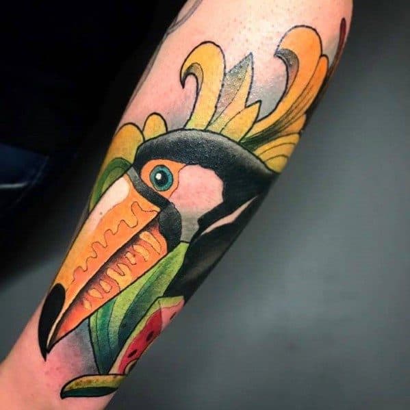 Colorful toucan tattoo on the forearm with exaggerated yellow and orange features, tropical leaves, and a watermelon slice accent.
