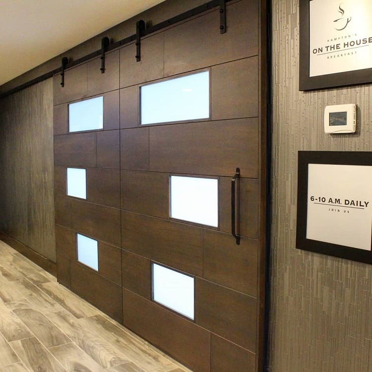 Track-mounted brown barn door with frosted glass panels in a hotel hallway with wood-patterned floors.