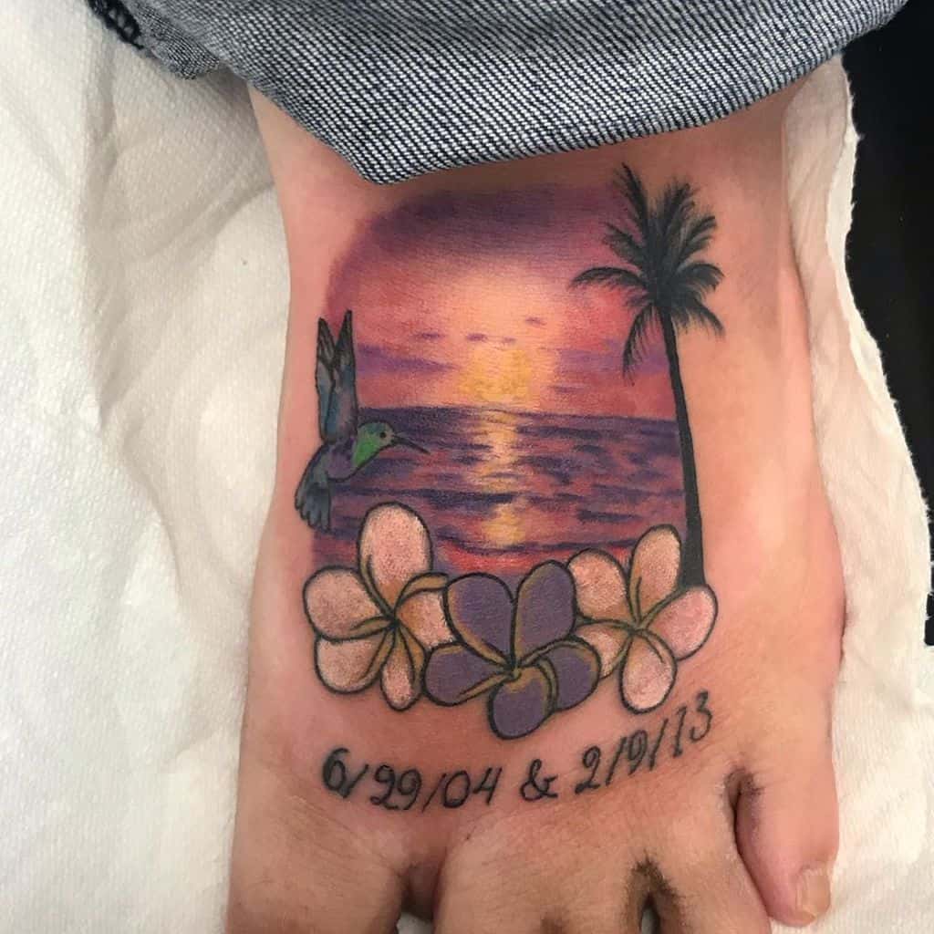 Foot tattoo featuring a sunset over the ocean, a palm tree, a hummingbird, plumeria flowers, and commemorative dates