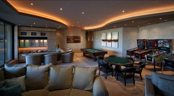 Luxurious game room with pool table, poker table, pinball machines, and modern seating under ambient lighting