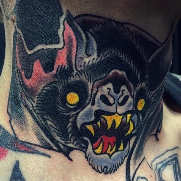 Intense traditional bat head tattoo with glowing yellow eyes, red mouth, sharp yellow fangs, and heavy black shading.
