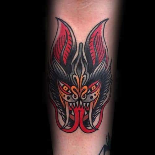 Traditional bat head tattoo with ornate red, orange, and black details, sharp fangs, and symmetrical tribal-inspired patterns.
