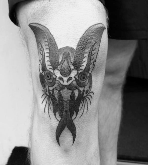 Blackwork bat head tattoo on the knee with symmetrical design, bold linework, and elongated tongue elements for a striking look.