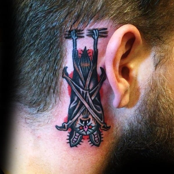 Small traditional bat tattoo behind the ear, hanging upside down from a branch with red background accents and bold black shading.