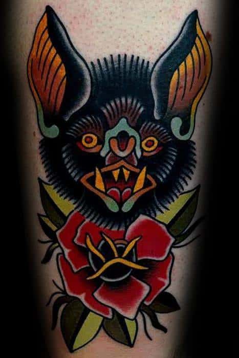 Traditional bat head tattoo with orange ears, yellow fangs, and a bold red rose below, framed by green leaves and thick black lines.