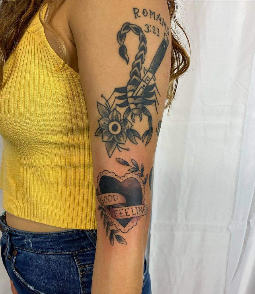 Tattooed woman's arm with a scorpion, dagger, flower, and heart-shaped banner reading "GOOD FEELING"