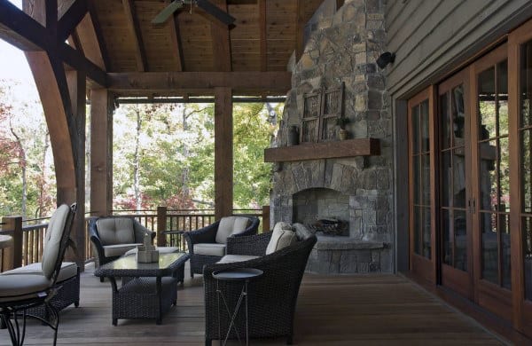 Cozy patio with wicker chairs, a glass table, and a stone fireplace under a wooden roof, overlooking a forest view