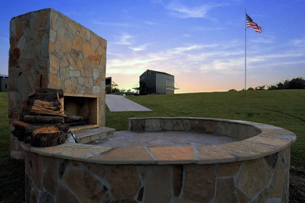 Stone fire pit with stacked wood beside it, American flag in the background, set against a twilight sky