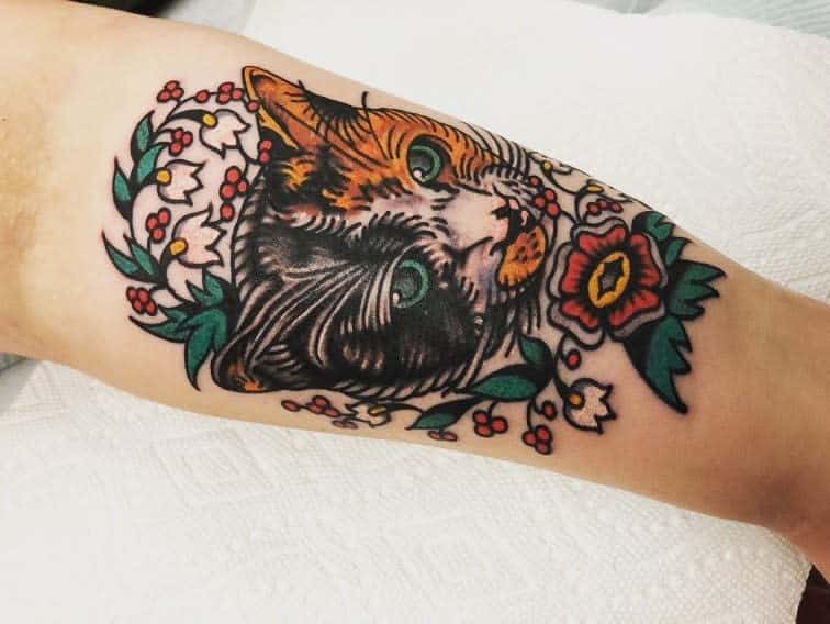 Vibrant neo-traditional tattoo of a calico cat with striking green eyes, surrounded by Lily of the Valley and red floral accents