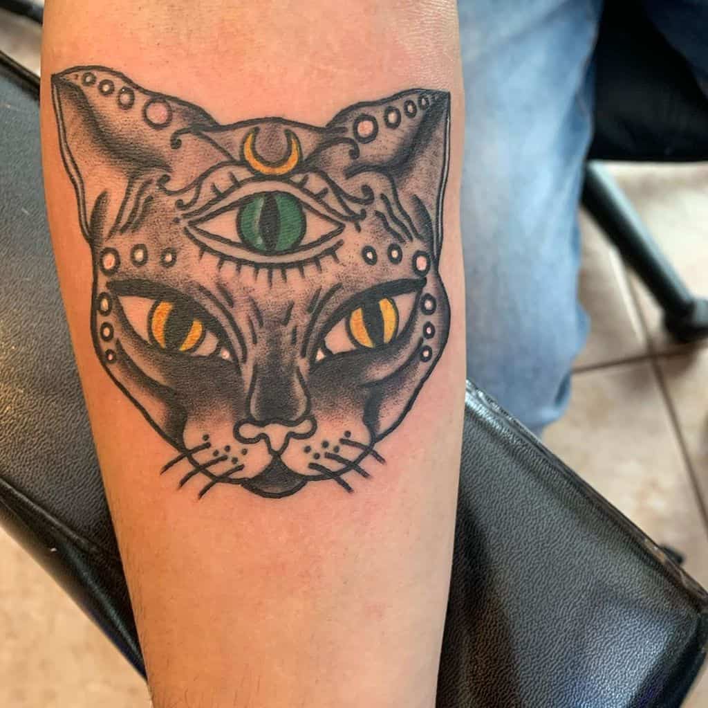 Tattoo of a mystical cat with a third eye, crescent moon, and intricate patterns, inked in bold black outlines with yellow and green accents on the forearm.