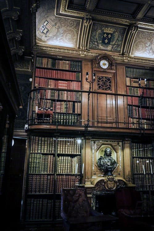 Opulent library with two levels of bookshelves, ornate ceiling, bust sculpture, and a large clock above the shelves