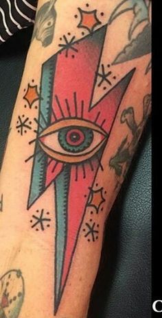 Tattoo of an all-seeing eye inside a bold red and blue lightning bolt, surrounded by radiating lines and sparkles, inked on the forearm.