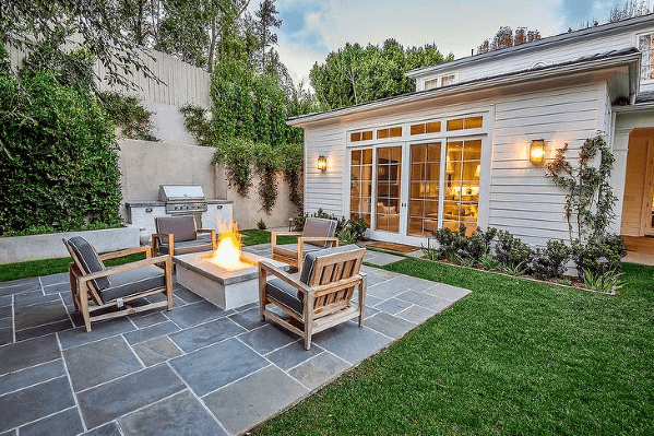 Backyard patio with a fire pit, wooden furniture, and built-in grill, surrounded by greenery and large windowed house