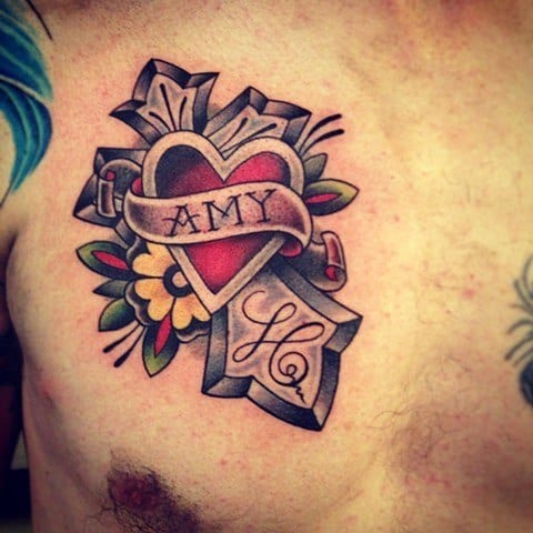 Traditional chest tattoo of a red heart with a banner reading "AMY," layered over a cross, with flowers, leaves, and bold shading.