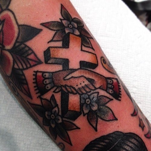 Traditional-style tattoo of a black and orange 3D cross with two hands shaking in front, surrounded by flowers and bold outlines.