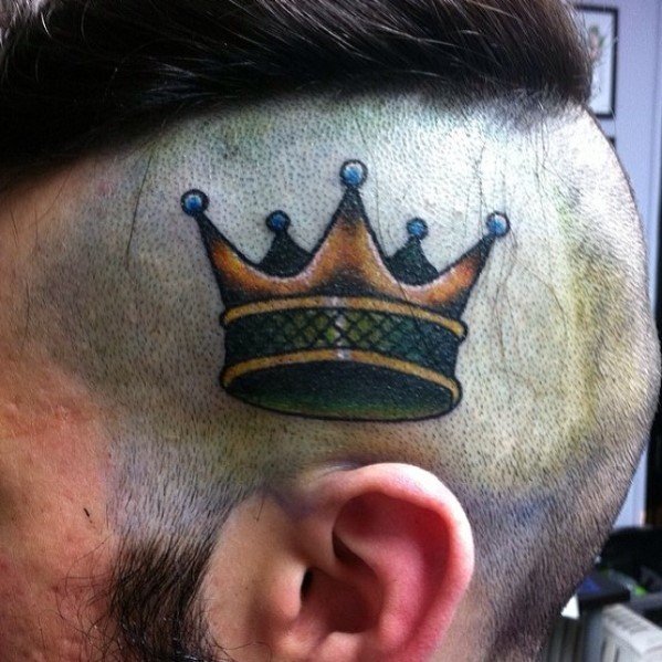 Realistic crown tattoo on the shaved side of the head, featuring gold and green tones with subtle shading and bold black outlines.