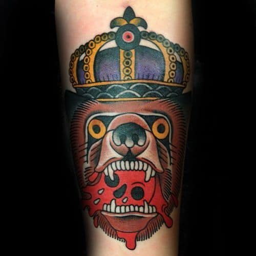 Bold traditional tattoo of a crowned bear with vivid yellow eyes, biting into a red skull, inked on the forearm with strong color contrast.