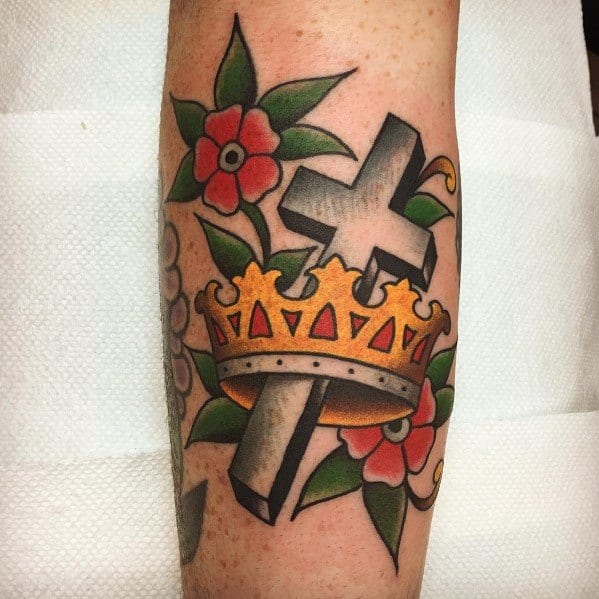 Traditional forearm tattoo featuring a shaded cross piercing through a gold crown, surrounded by bold red flowers and green leaves.