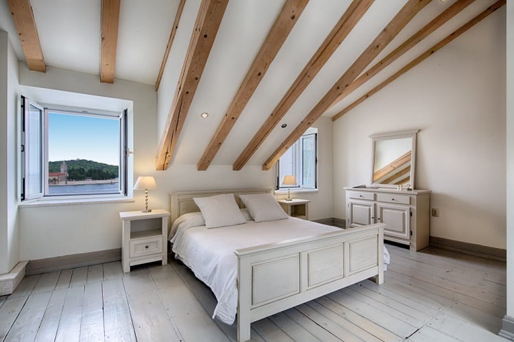 Cozy bedroom with sloped wooden ceiling beams, a double bed, and a dresser by an open window overlooking greenery