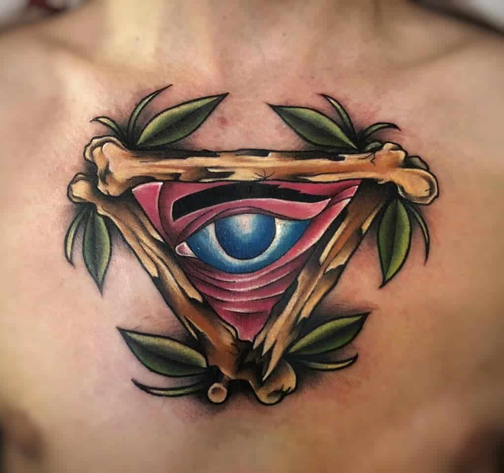 Tattoo of an all-seeing eye inside a triangle made of bones, surrounded by green leaves, inked in vibrant colors on the chest.