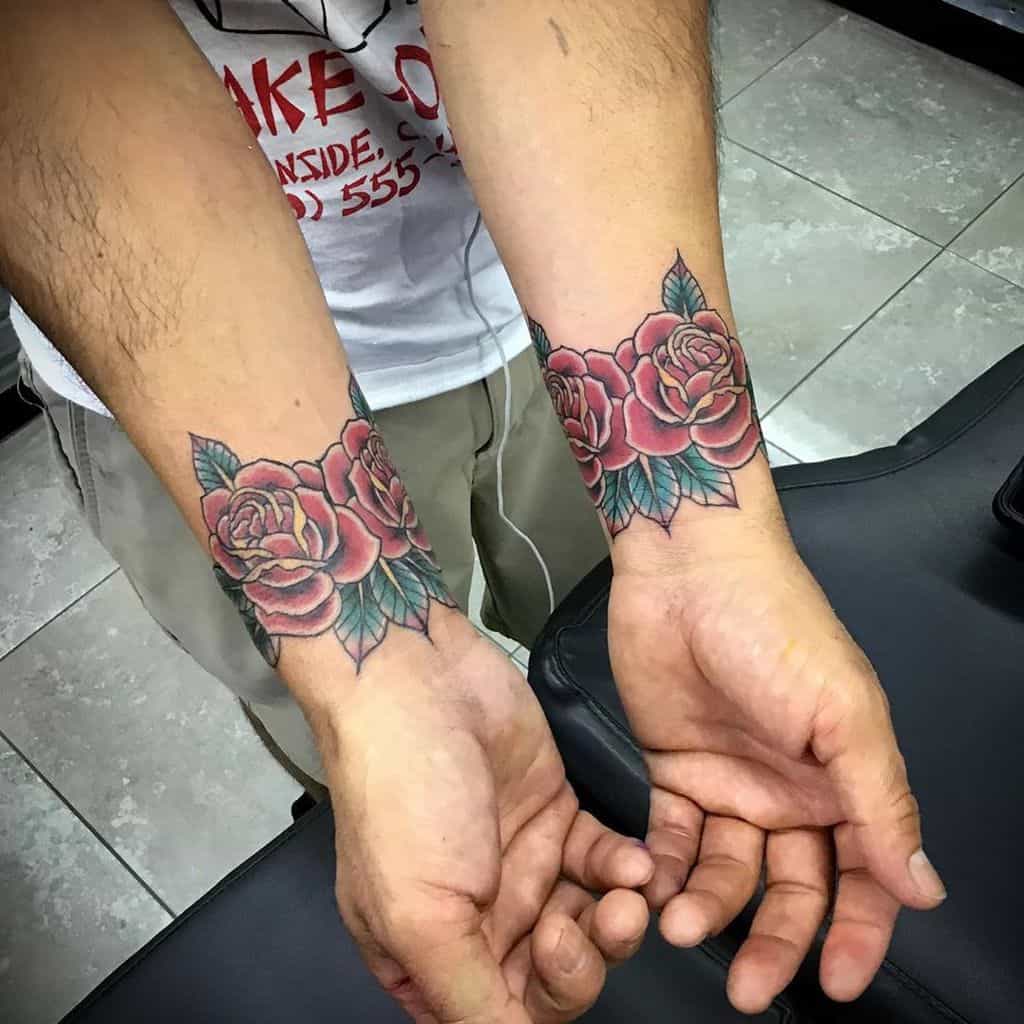 Matching traditional style rose tattoos with vibrant red petals and green leaves, placed on both wrists