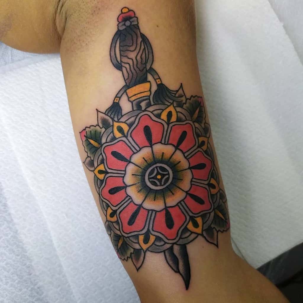 Colorful flower tattoo with a dagger design on an arm, featuring red petals and green leaves