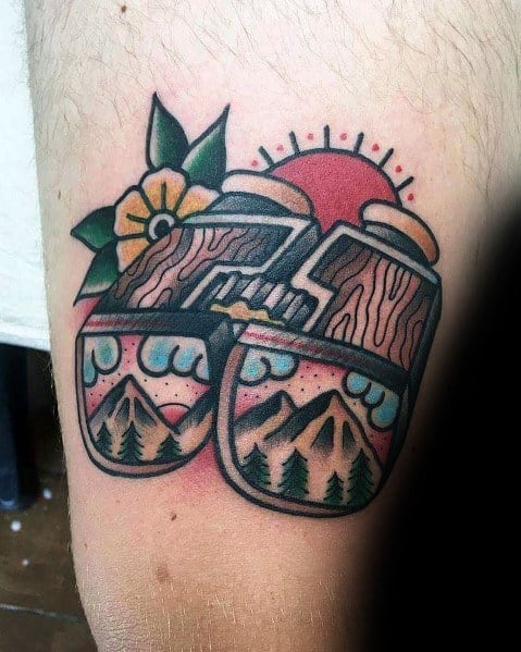 Colorful tattoo of binoculars reflecting a scenic mountain view, with wood texture and floral details, symbolizing adventure and nature.