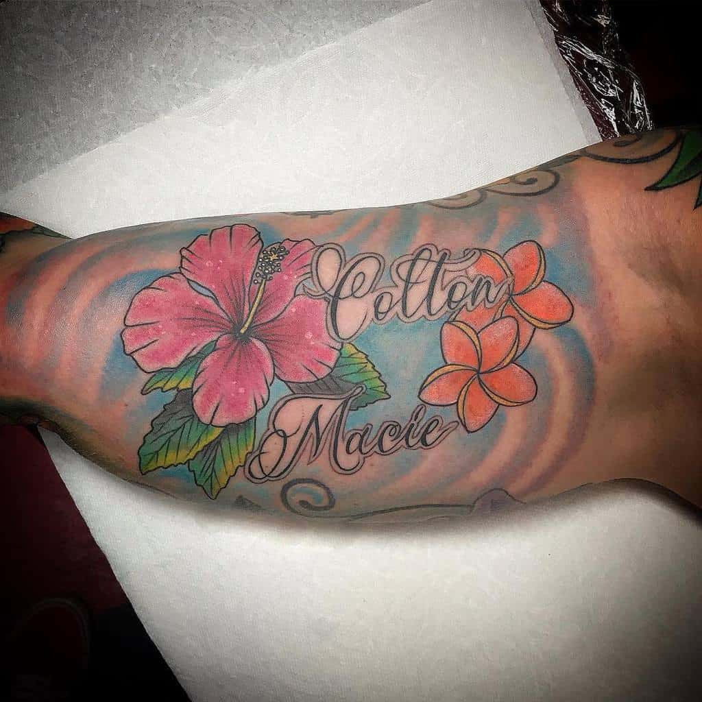 Forearm tattoo featuring a pink hibiscus and orange plumeria flowers with scripted names 'Cotton' and 'Macie' against a blue swirl background