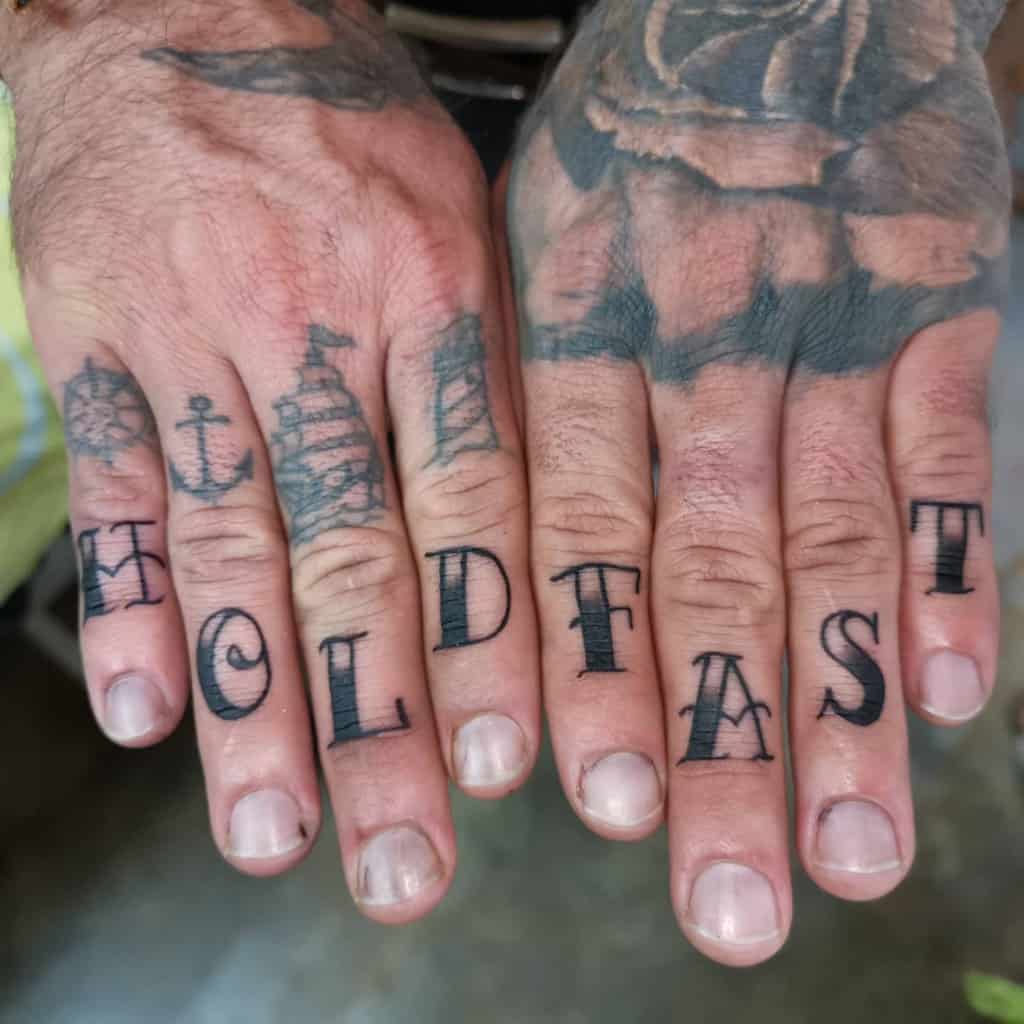 Hands with "HOLD FAST" tattoos on fingers and nautical symbols on the knuckles