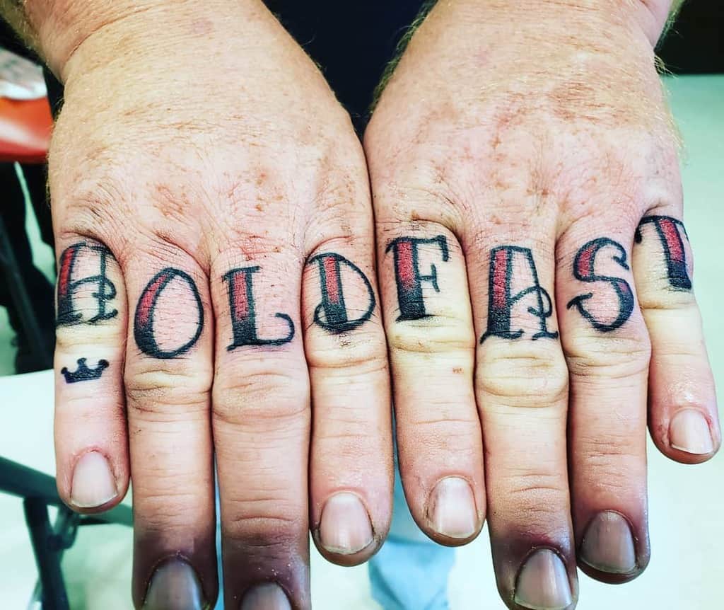 Hands with "HOLD FAST" tattooed across the fingers, each letter in black and red ink