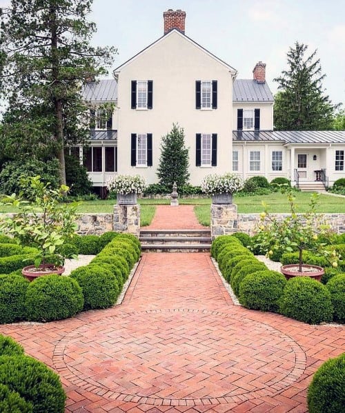 Classic brick walkway with a circular pattern, surrounded by manicured boxwood hedges, leading to a traditional home with a charming garden