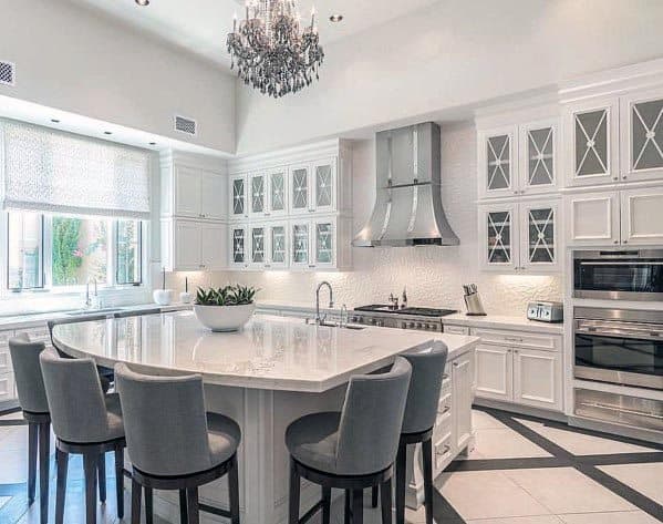 White kitchen with glass cabinet doors, marble island, grey bar stools, and chandelier.