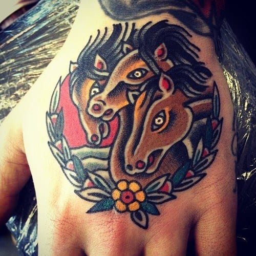 Traditional hand tattoo featuring three horse heads with red accents, framed by a floral wreath and a sunset-style background.