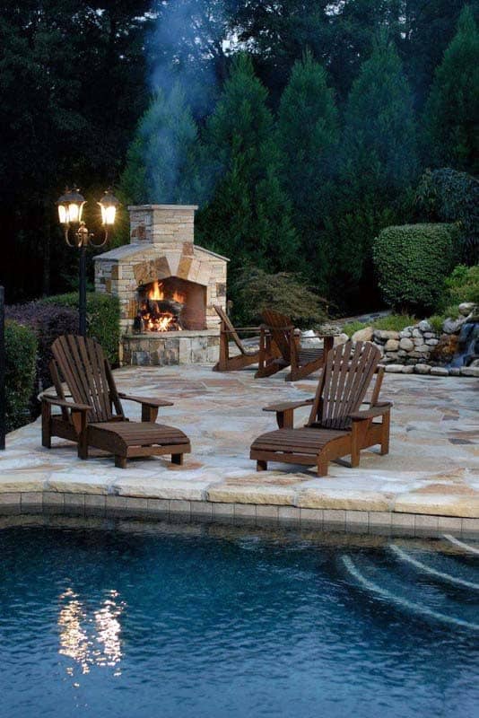 Poolside patio with Adirondack chairs, a lit stone fireplace, and lanterns surrounded by trees and shrubs