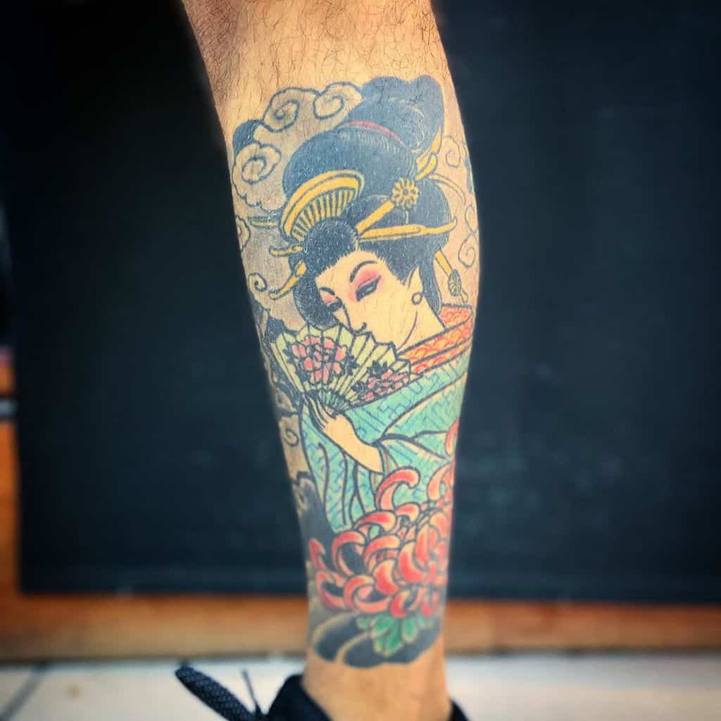 Traditional Japanese-style leg tattoo of a Geisha in a blue kimono, holding a fan, with floral patterns, swirling clouds, and ornate hairpieces.