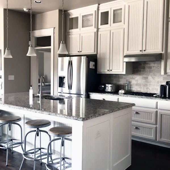 Classic kitchen with white cabinetry, textured stone backsplash, and a granite island featuring industrial-style bar stools
