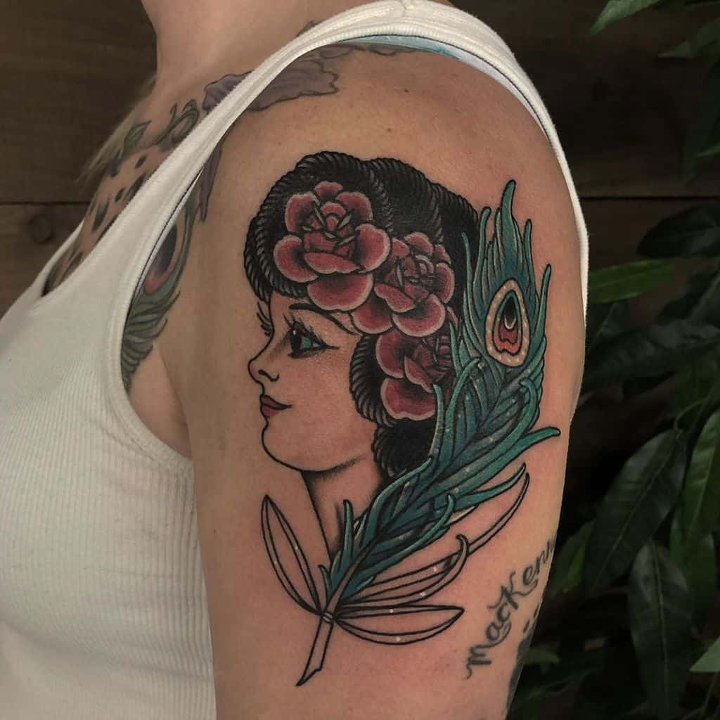 Traditional-style tattoo on the upper arm featuring a woman with flowers in her hair, a detailed peacock feather, and bold black outlines.