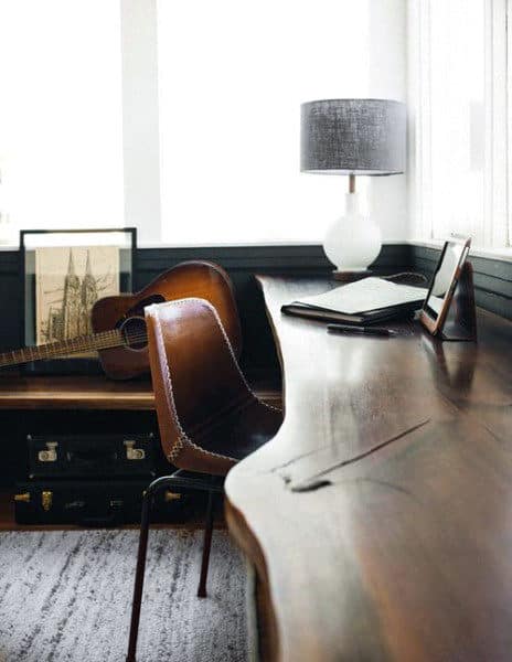 A cozy workspace with a wooden desk, leather chair, guitar, lamp, and artwork; natural light fills the room