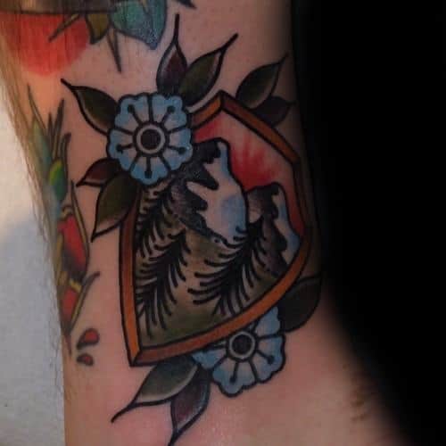Traditional tattoo of a mountain landscape with pine trees inside a shield-shaped frame, accented by blue flowers and green leaves.