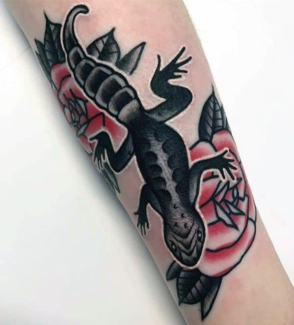 Bold black gecko tattoo on forearm with traditional style, crawling over two shaded pink roses and dark green leaves.