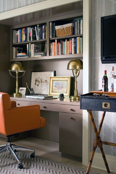 Home office with an orange chair, bookshelf, two brass lamps, and a desk with framed pictures and books