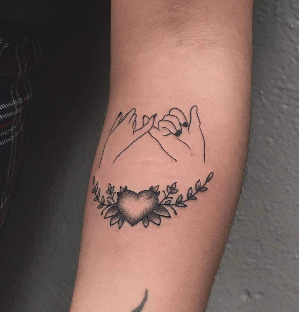 Tattoo of two hands making a pinky promise, with a heart and leaves beneath them, on an arm